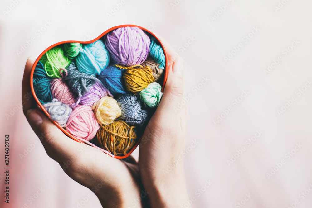 Heart-shaped box with knitted multi-colored skeins of yarn in hands on wooden background. Crochet and knitting. Women's working space. 