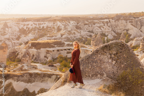 Beautiful traveling woman wearing authentic boho chic style red dress and hat looking to sunrise sky in Cappadocia valley. Travel and wanderlust concept. Copy space background.