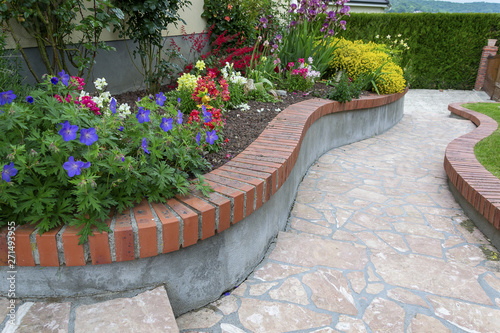 Allée fleurie (ancolie, mufliers, gueule de loup, genet, iris) avec un sol en marbre rose du portugal sur muret courbe surmonté de brique terre de France