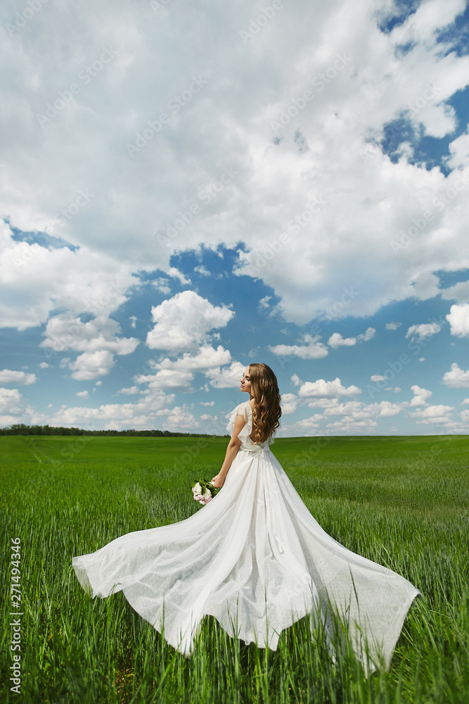 Beautiful young model woman with wedding hairstyle in white long dress posing in a green field in the summer sunny day Beautiful young model woman with wedding hairstyle in white long dress posing in a green field in the summer sunny day