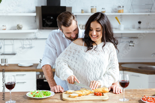 happy couple standing together at kitchen while woman cutting bread on chopping board
