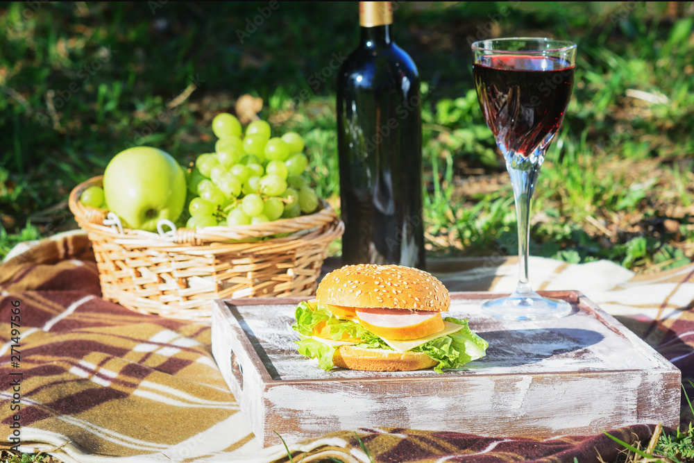 Summertime Picnic Lunch at Green Sunny Meadow. Wicker Basket Full of Fresh Grape and Apple. Bottle of Red Wine Ham and Cheese Burger, Wineglass Served on Wooden Tray. Pleasant Outdoors Dinner