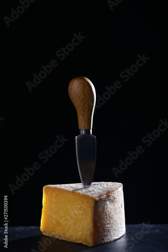A large piece of hard cheese with a knife for cutting cheese on a black background. Isolated object. Craft cheese concept. Still life concept.