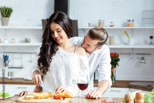handsome man cutting bread on chopping board and kissing girlfriend shoulder