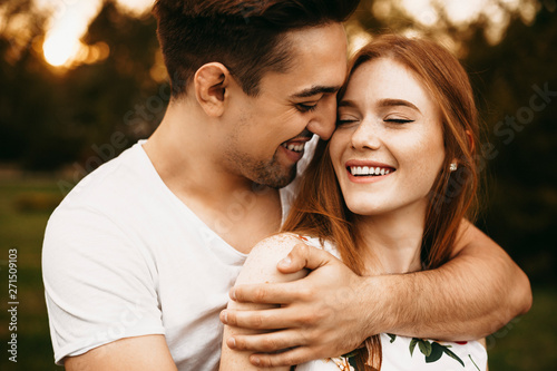 Slika na platnu Portrait of a beautiful red hair woman with freckles smiling with eyes closed while being embraced by her boyfriend outside while dating against sunset