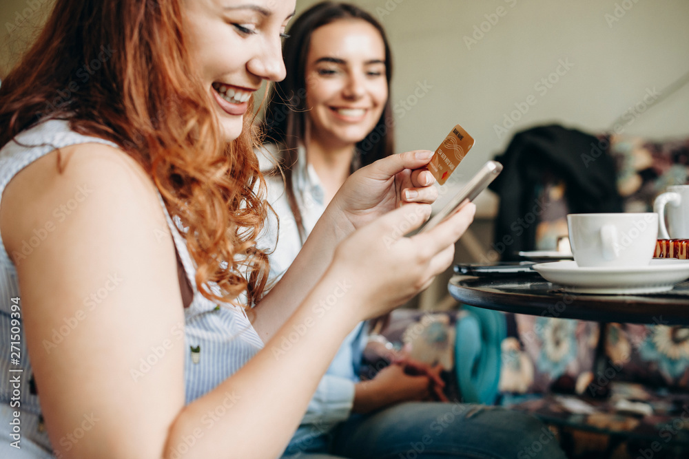 Side view of a female hands using a credit gold card and a smartphone for online banking while sitting a desk in coffee shop with a female friend smiling. Side view of a female hands using a credit gold card and a smartphone for online banking while sitting a desk in coffee shop with a female friend smiling.