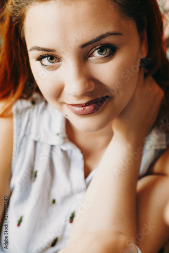 Close up portrait of a beautiful plus size women with red hair looking at the camera smiling while holding a hand on her neck.