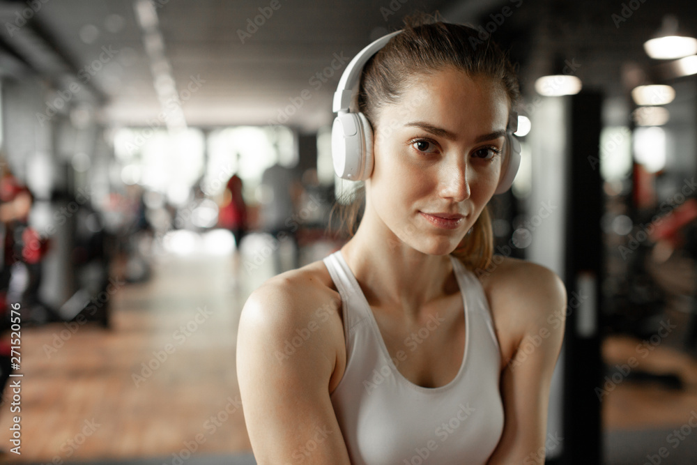 Close up portrait of young pretty european fitness woman at the gym and listen music in headphones. Breaking relax while exercise workout. Concept of health and sport lifestyle. Athletic Body..