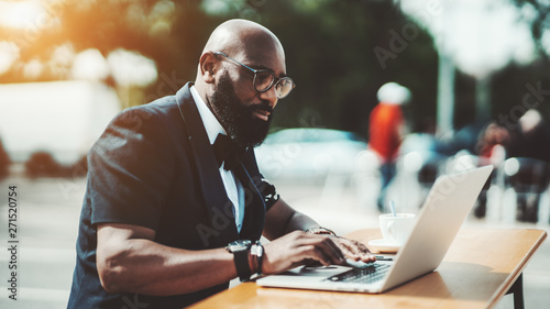 A strong adult bold African man entrepreneur in eyeglasses is working on the laptop in an outdoor cafe; a bearded businessman in a street bar with his netbook and a cup of delicious coffee