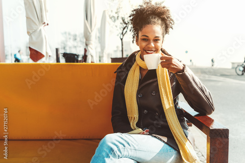 A Dazzling young happy African-American female is drinking a delicious coffee drink in an outdoor bar; smiling Brazilian girl in demi-season coat and scarf is drinking tea in a cafe outdoors