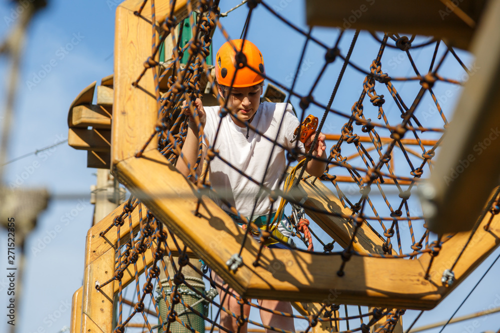 Fototapeta premium Child in forest adventure park. Kid in orange helmet and white t shirt climbs on high rope trail. Agility skills and climbing outdoor amusement center for children. young boy plays outdoors