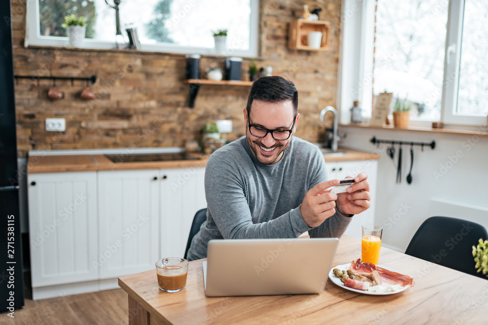 Smiling handsome man with credit card looking at laptop at the kitchen table with breakfast. Smiling handsome man with credit card looking at laptop at the kitchen table with breakfast.
