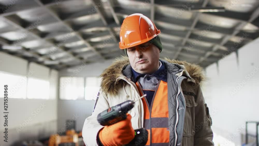 man in a hard hat and overalls is standing in the middle of the hangar and cheking a drill at the camera man in a hard hat and overalls is standing in the middle of the hangar and cheking a drill at the camera