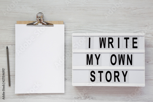 'I write my own story' words on a lightbox, clipboard with blank sheet of paper on a white wooden background, top view. From above, overhead, flat lay.