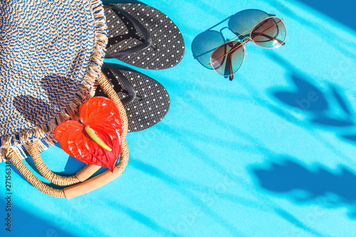 Summer accessories on sunny beach, trendy feminine woven straw bag with red flower, flip flops and lovely shadows on blue background. Summer exotic travel and vacation concept. Flat lay with copyspace photo