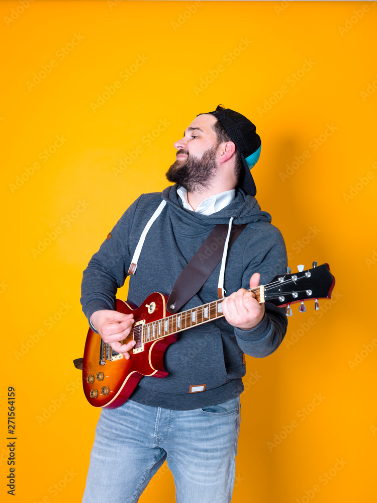 Obraz premium man with black hair and beard, wearing grey hoodie playing the electric guitar in front of a yellow background