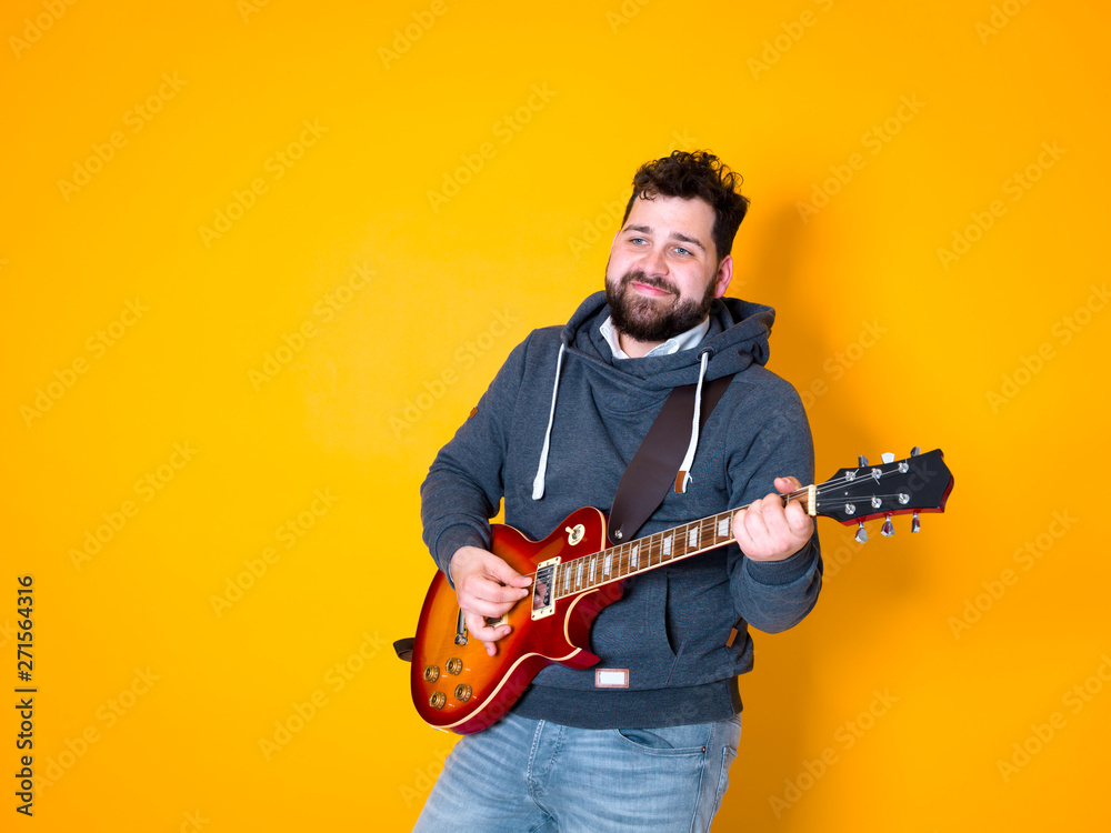 Obraz premium man with black hair and beard, wearing grey hoodie playing the electric guitar in front of a yellow background