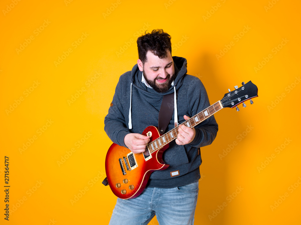 Obraz premium man with black hair and beard, wearing grey hoodie playing the electric guitar in front of a yellow background