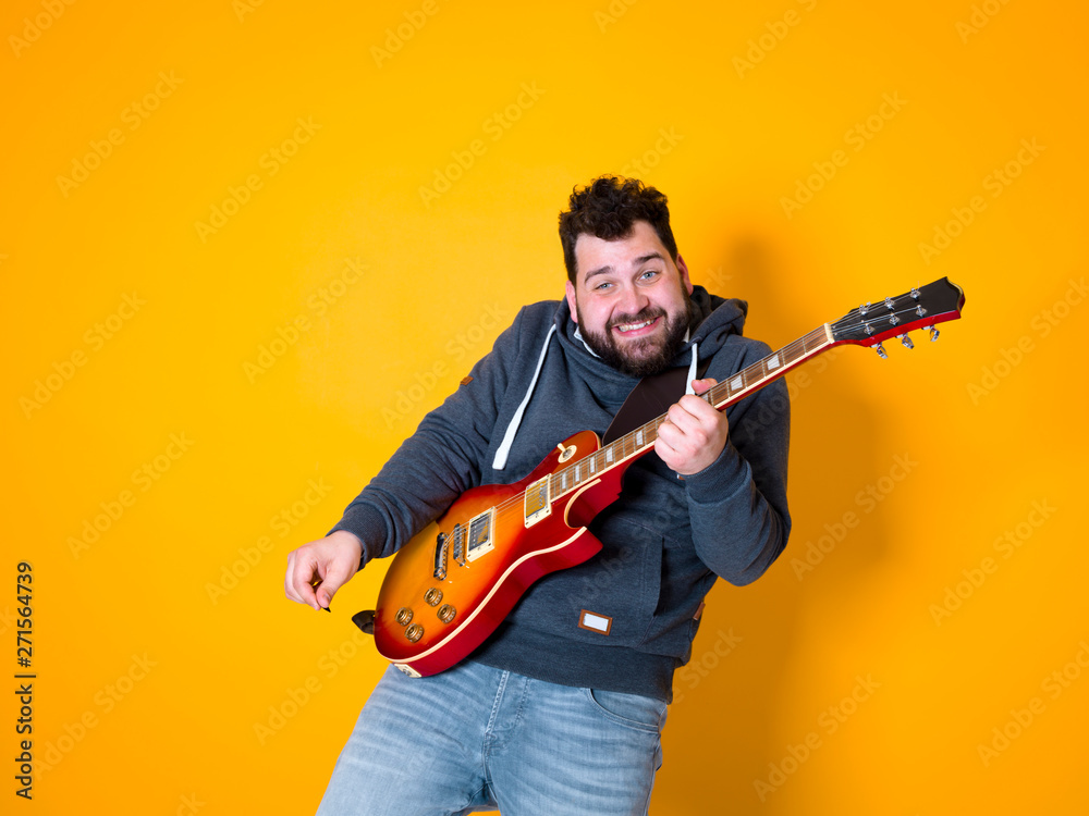 man with black hair and beard, wearing grey hoodie playing the electric guitar in front of a yellow background man with black hair and beard, wearing grey hoodie playing the electric guitar in front of a yellow background