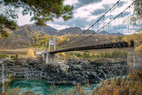 Road bridge over turquoise Katun river in Altai Mountains. Russian autumn in Siberia. Incredible landscape with a metal bridge over the water, rocky coast, hill ridge in the background, storm clouds.