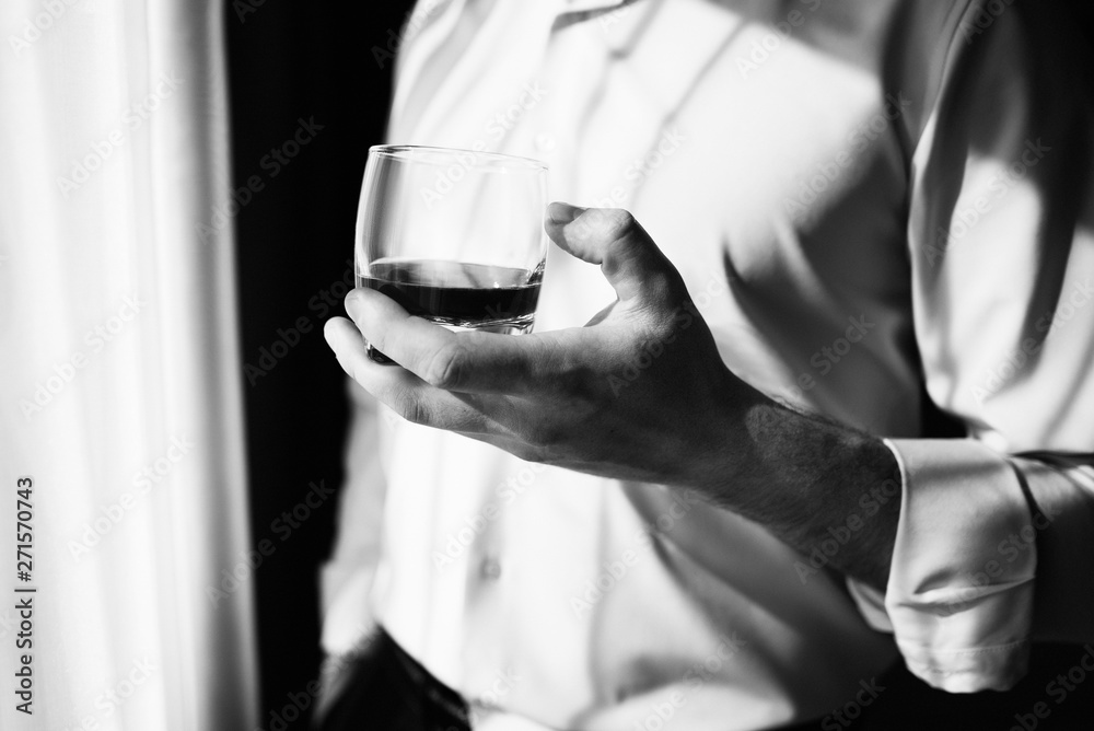 Young man with glass on whiskey near window. Man drinking whiskey at the window. Drink whiskey, cognac, close-up. Hand with a glass of alcohol. Black and white photo