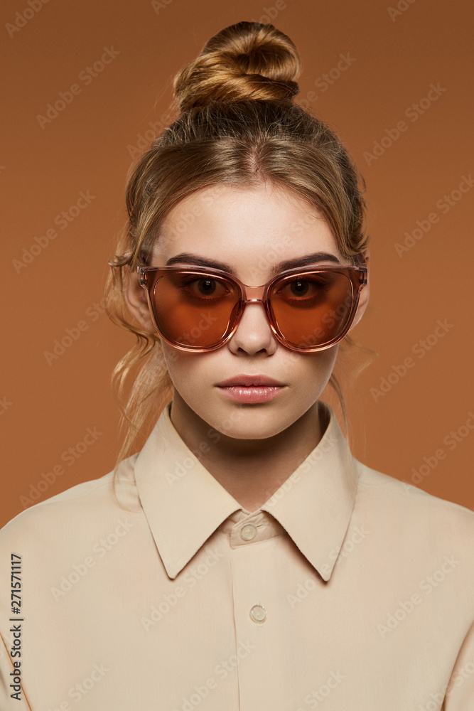 Cropped front view shot of blonde lady, wearing shirt. The girl with bun and wavy hair locks in butterfly sunglasses with light brown rim and lenses, is looking at camera over the brown background.