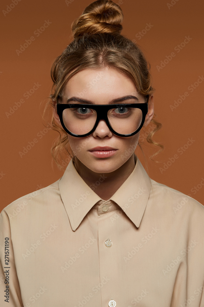 Cropped front view shot of lady, wearing shirt. The girl with bun and wavy hair locks in wrap-shaped glasses with black rim and transparent lenses. The woman is looking at camera on brown background.