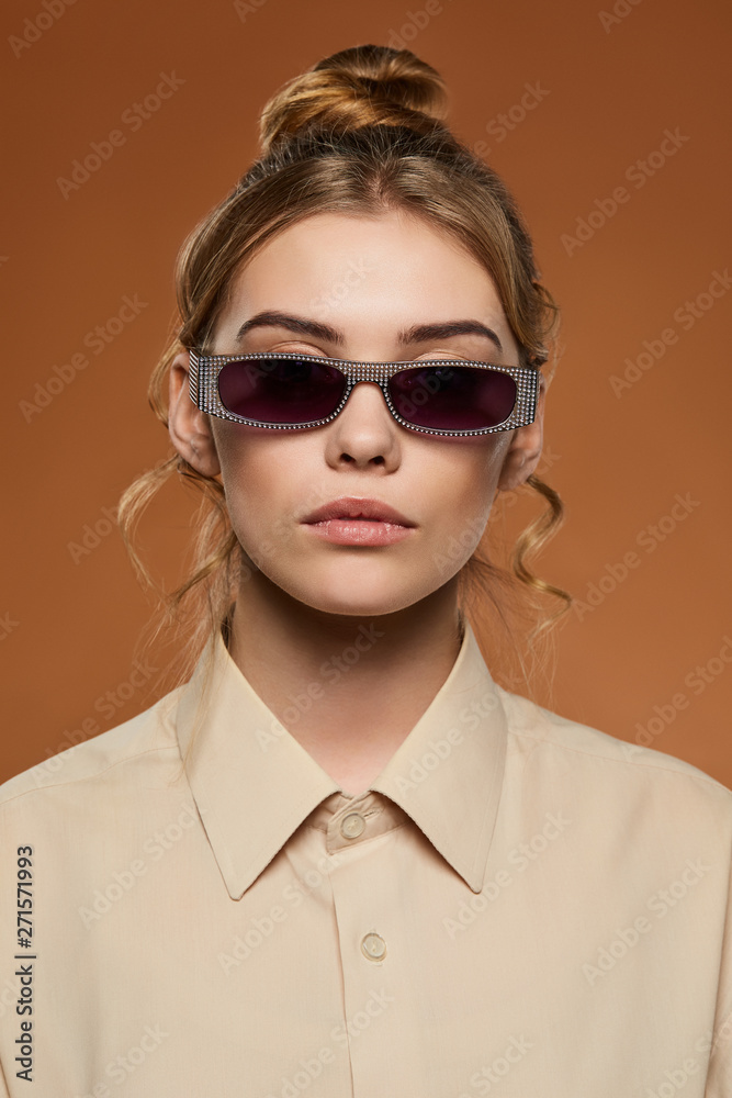 Cropped front view shot of blonde lady, wearing shirt. The girl with bun and wavy hair locks in wine red rectangle sunglasses with silver toreutics on rim, is looking at camera on sandy background.