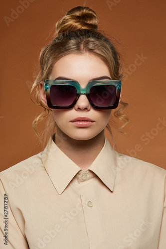 Cropped front view shot of blonde lady, wearing shirt. The girl with bun and wavy hair locks in square-shaped sunglasses with gradient rim and black lenses, is looking at camera on brown background. © RedUmbrella&Donkey