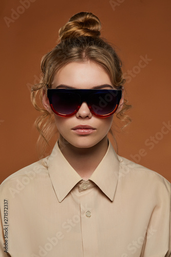 Cropped front view shot of woman, wearing shirt. The girl with bun and wavy hair locks in wrap sunshades with dark red gradient rim and lenses. The lady is looking at the camera on brown background.