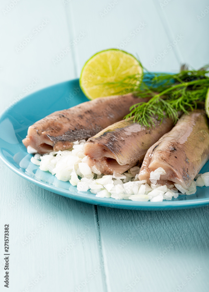 Traditional dutch food freshly salted herring fish with onion called hollandse nieuwe on turquoise plate and wooden background. European food concept with copy space. Traditional dutch food freshly salted herring fish with onion called hollandse nieuwe on turquoise plate and wooden background. European food concept with copy space.