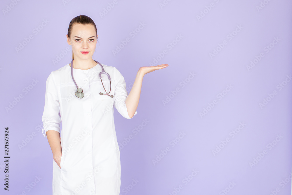 Young female blonde doctor with stethoscope on her neck points her hand at empty space for text on purple background. Doctor looks at camera Young female blonde doctor with stethoscope on her neck points her hand at empty space for text on purple background. Doctor looks at camera