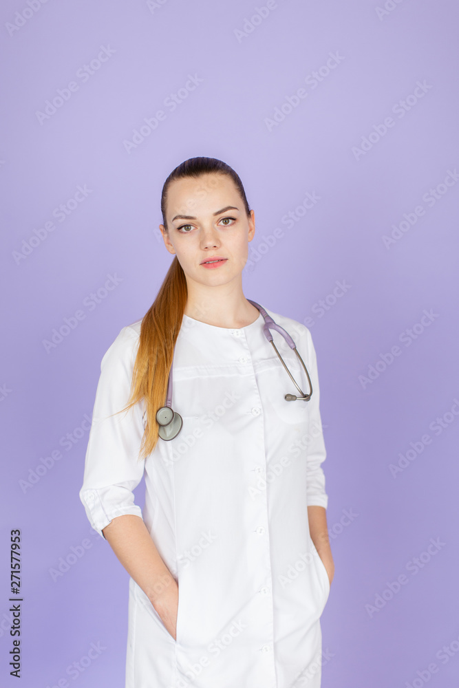 Young female blonde doctor with her hands in pockets with stethoscope on her neck on purple background. Doctor looks at camera Young female blonde doctor with her hands in pockets with stethoscope on her neck on purple background. Doctor looks at camera