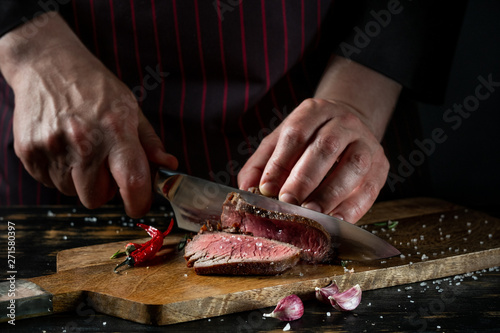 Slicing juicy beef steak by knife in chef hands closeup. Food cooking concept. Dark black background copy space.