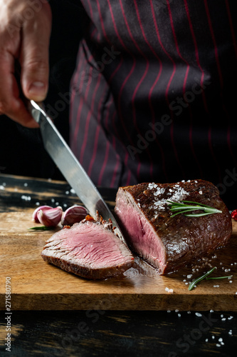 Slicing juicy beef steak by knife in chef hands closeup. Food cooking concept. Dark black background copy space.