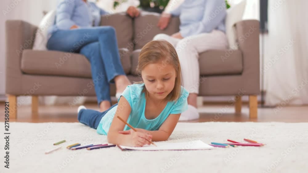 family, childhood and leisure concept - girl drawing in sketchbook by crayons lying on floor and adults sitting on sofa at home