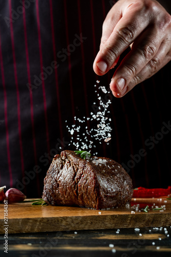 Chef hands cooking meat steak and adding salt and pepper on black copy space background for menu restaurant or recipe text.