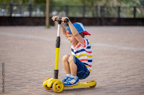 Two boys, one in a cap, the other in a hood, roll on a scooters pushing off a road with smiles on their face around square for walks and rest on  summer