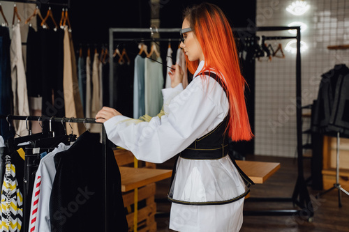 Attractive Redhead Woman in Showroom Side View. Young Beauty Choosing Trendy Apparel. Pretty Caucasian Redhead Looking at Rack with Designer Clothes Collection. Commercial Catalog Shoot