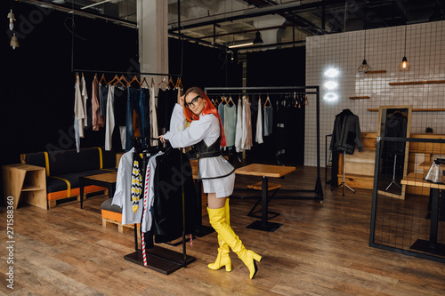 Beautiful Woman Posing in Fashion Garment Boutique. Stylish Caucasian Redhead Girl Lean on Rail with Clothes. Model Wearing Glasses and Trendy Look. Modern Showroom Inside Interior