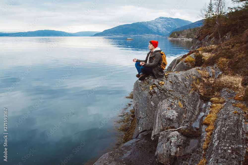 Traveler man in a meditative position sitting on a rocky shore on the background of a mountain and a lake. Space for your text message or promotional content.