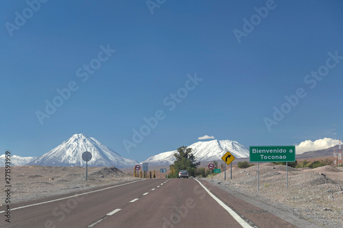 Long straight road with yellow road sign leading towards a snow capped mountain in Atacama desert, Chile