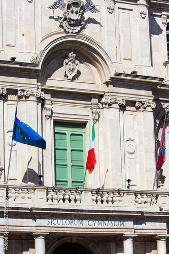 Detail vertical photography capturing front side exterior facade of historical university in Catania, Sicily, Italy. Baroque architecture style, green window and waving Italian flag and flag of EU