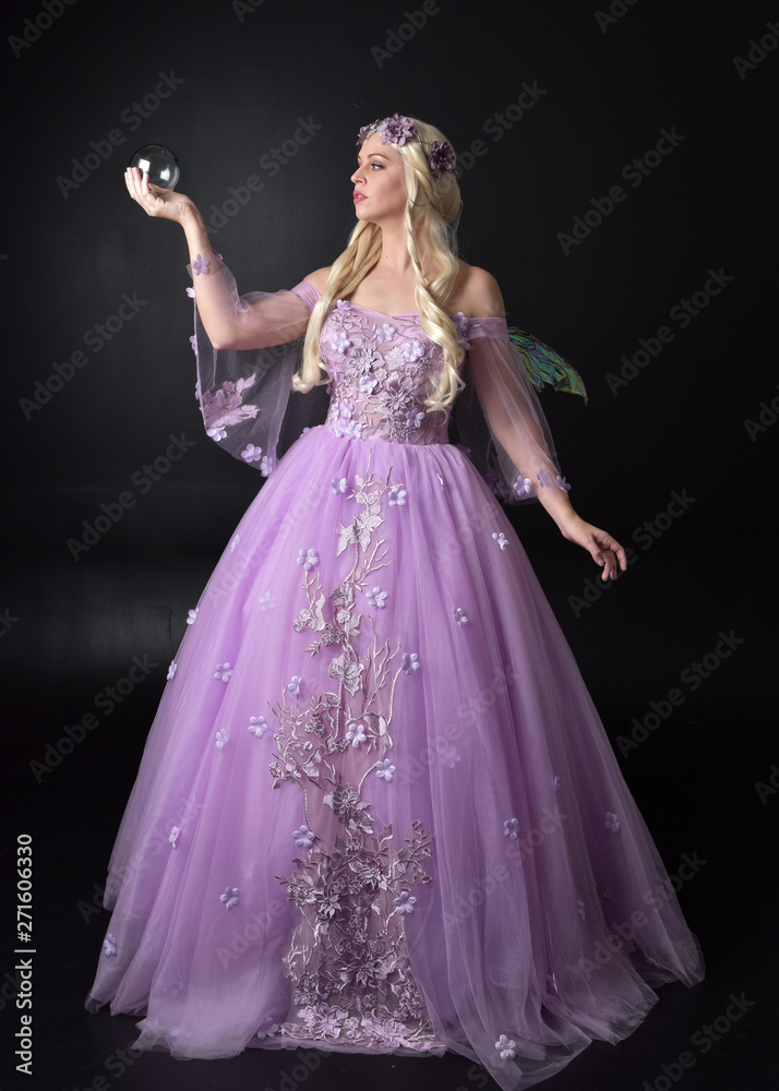 full length portrait of a blonde girl wearing a fantasy fairy inspired costume,  long purple ball gown with fairy wings,  standing pose holding a crystal ball on a dark studio background.