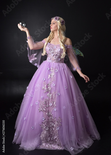 full length portrait of a blonde girl wearing a fantasy fairy inspired costume, long purple ball gown with fairy wings, standing pose holding a crystal ball on a dark studio background.