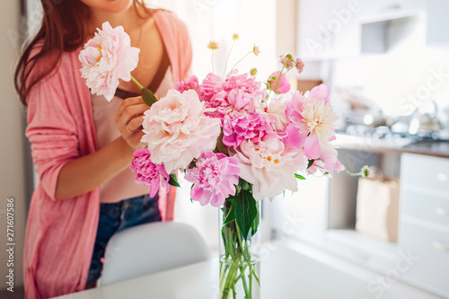 Woman puts peonies flowers in vase. Housewife taking care of coziness and decor on kitchen. Composing bouquet.