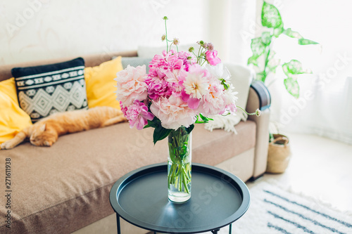 Flat interior of living room decorated with bouquet of peonies, basket, plants and carpet. Cat sleeping on couch