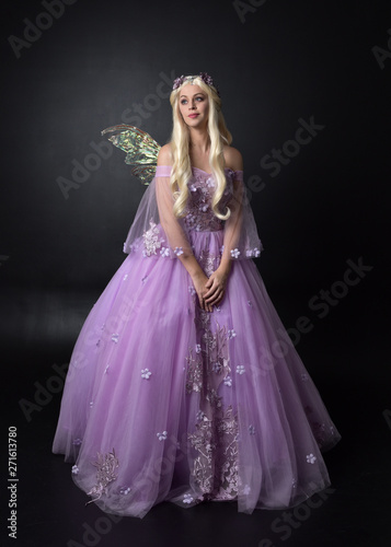 full length portrait of a blonde girl wearing a fantasy fairy inspired costume,  long purple ball gown with fairy wings,   standing pose on a dark studio background.