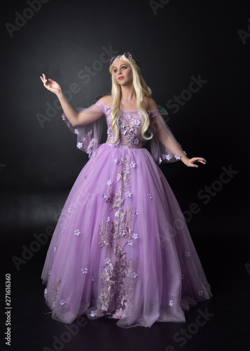 full length portrait of a blonde girl wearing a fantasy fairy inspired costume, long purple ball gown with fairy wings, standing pose on a dark studio background.