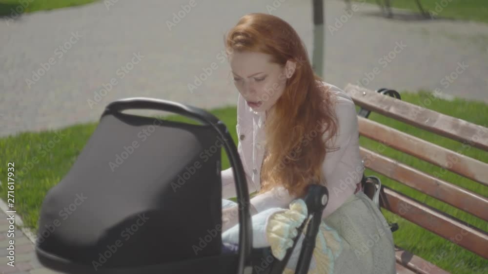 Portrait red-haired woman playing with her child sitting on the bench. The stroller standing near. The lady enjoying the sunny day with her baby outdoors. Young mother with a kid. Happy family Portrait red-haired woman playing with her child sitting on the bench. The stroller standing near. The lady enjoying the sunny day with her baby outdoors. Young mother with a kid. Happy family